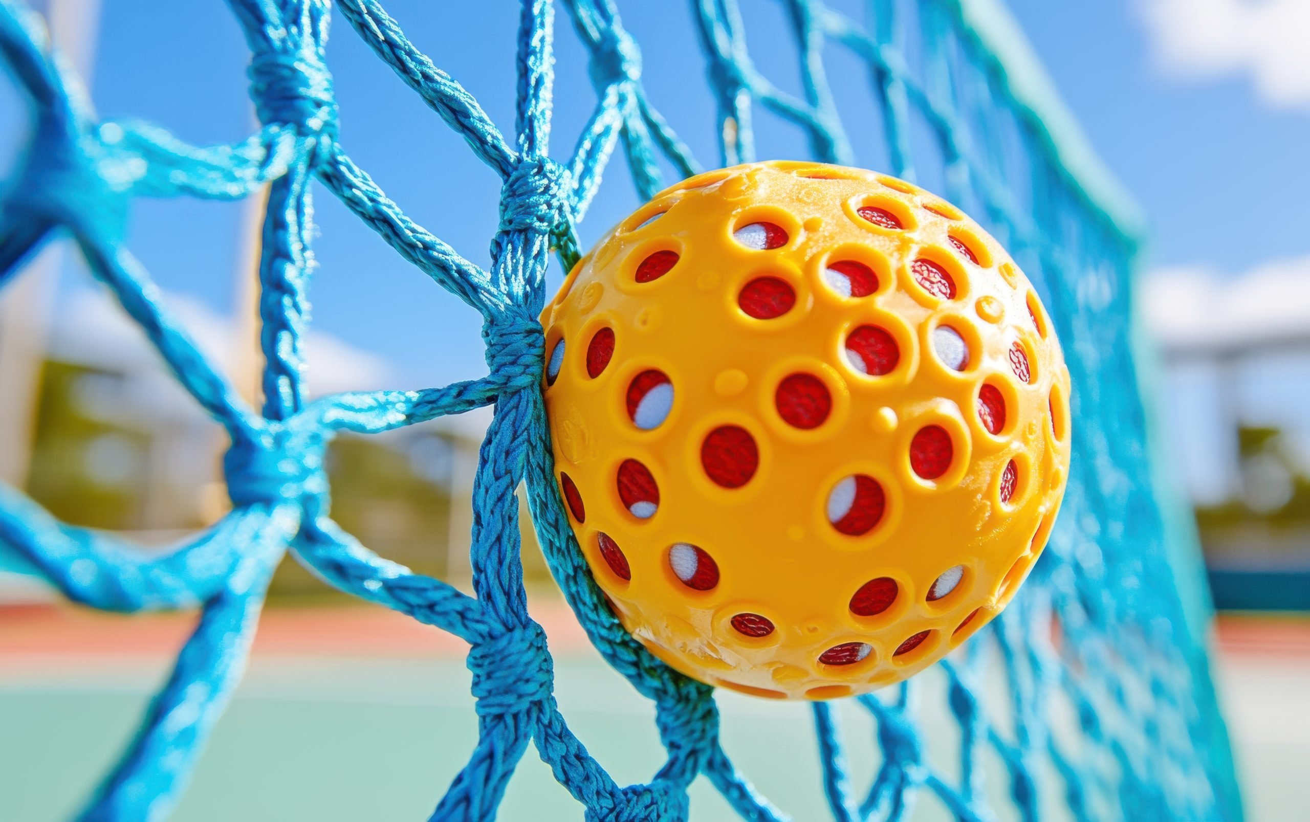 Close-up view of a pickleball net with a yellow ball stuck in the mesh during daylight on a sunny court
