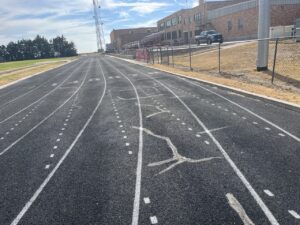 Weathered running track with faded lane markings and visible surface cracks near a school.
