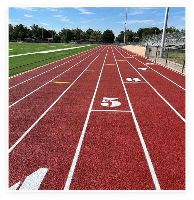 Red running track with white lane numbers and lines beside green turf field under clear blue sky