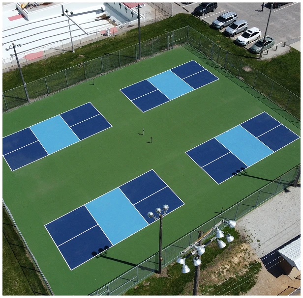 Multi-court pickleball facility construction and investment overview by Pro Track & Tennis Aerial view of four blue pickleball courts on a green surface surrounded by fencing at a sports facility
