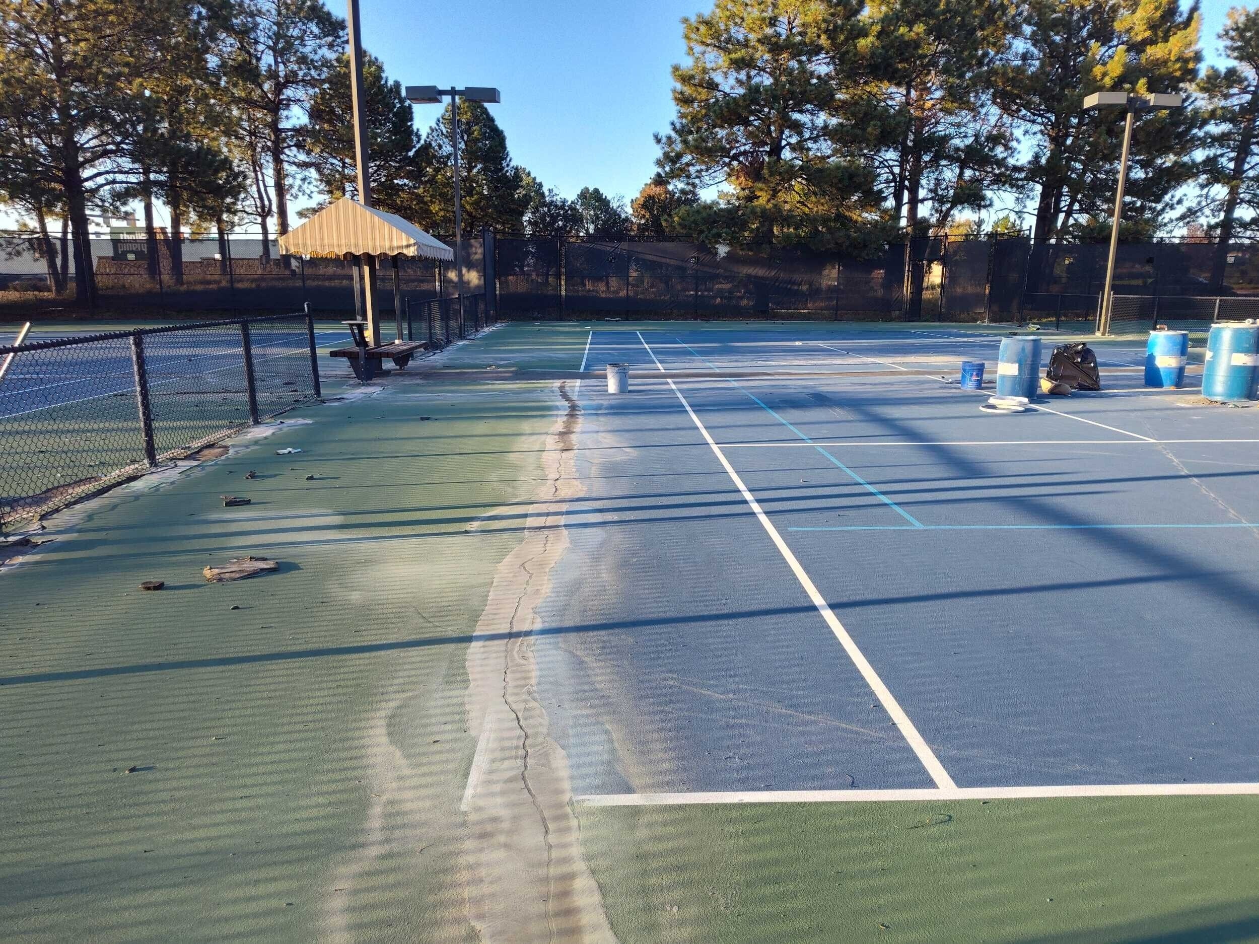 Tennis court undergoing resurfacing with visible cracks and base repairs before new UV-resistant coating.