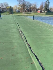 Long crack running along the surface of an aging tennis court, showing separation in the pavement.