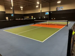 Indoor pickleball court with green and orange surface, portable net, and wall-mounted scoreboard