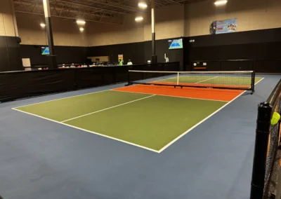 Indoor pickleball court with green and orange surface, portable net, and wall-mounted scoreboard