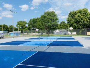 Blue pickleball courts at Imperial City with fencing, trees, and a nearby parking area.
