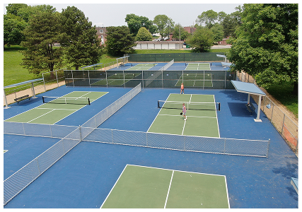 Aerial view of multi-court tennis facility with blue and green playing surfaces surrounded by fencing and trees