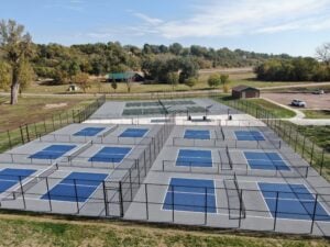 Aerial view of multi-court pickleball facility with fencing, surrounded by trees and open field