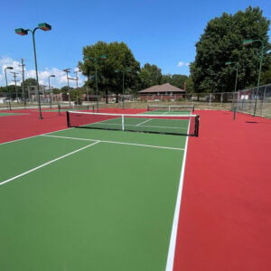 Green and red pickleball court with net, surrounded by trees and lighting fixtures